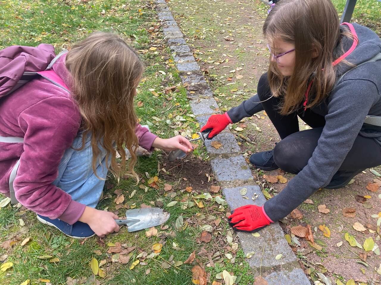 zwei Mädchen hocken vor einem kleinen Loch. Eine hält eine Blumenzwiebel in der Hand, die andere lächelt.