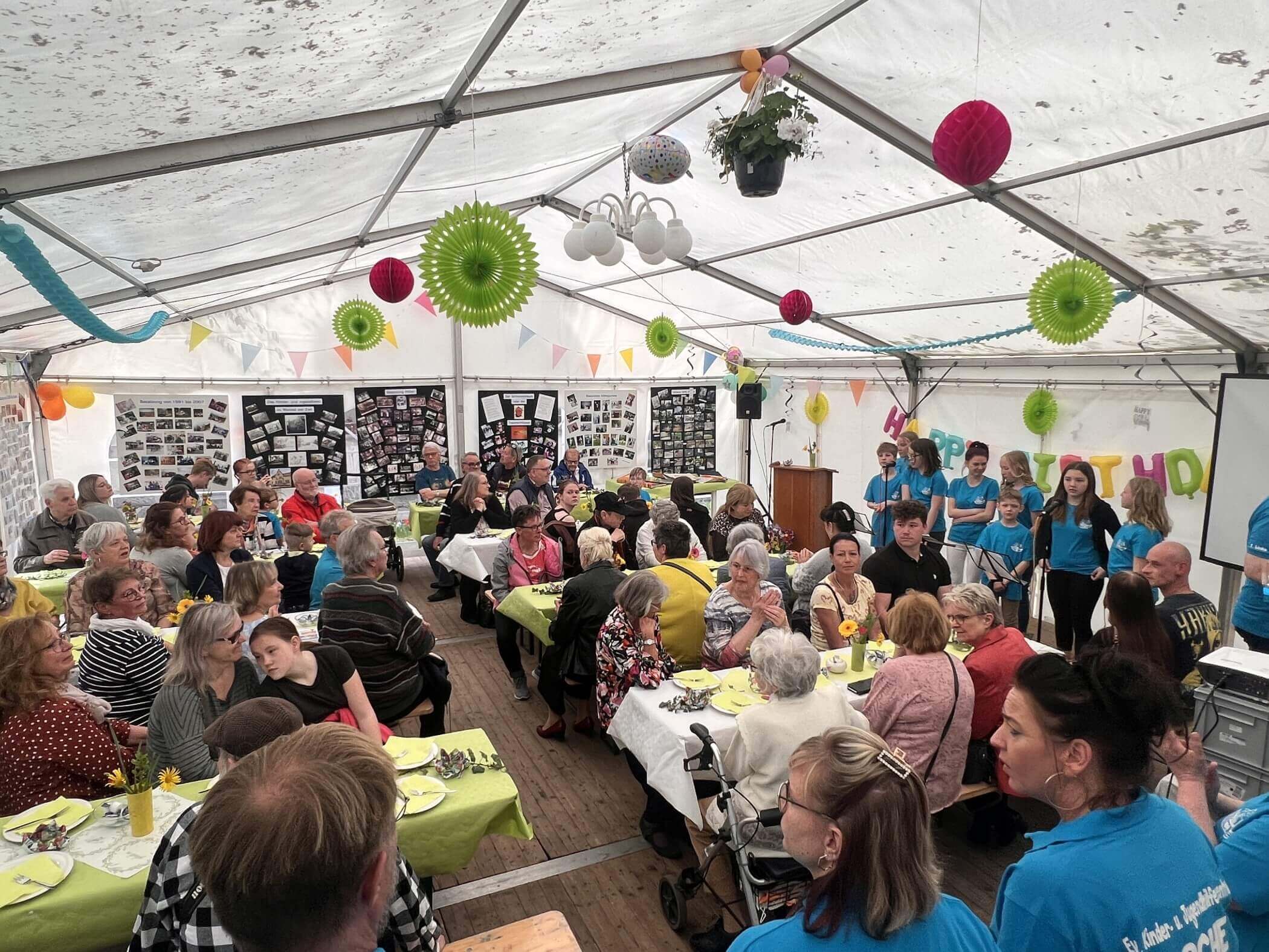 Blick ins Festzelt. Viele Menschen sitzen an Tischen, Ballons und Girlanden hängen von der Decke. Ein Chor singt.