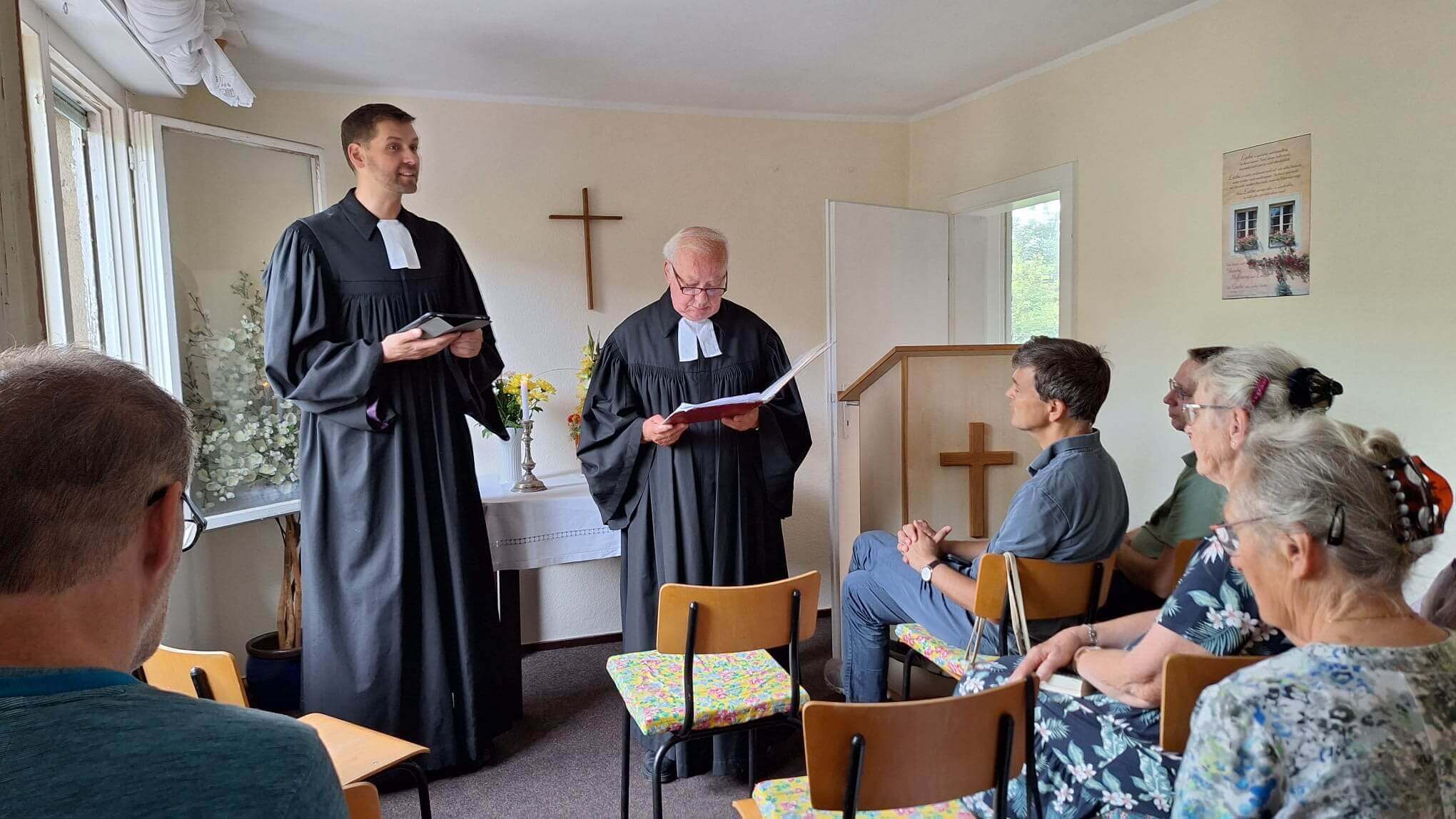 Blick in einen kleinen Raum mit Kreuz an der Wand. Menschen sind von hinten zu sehen. Zwei Personen im Talar stehen vorn.