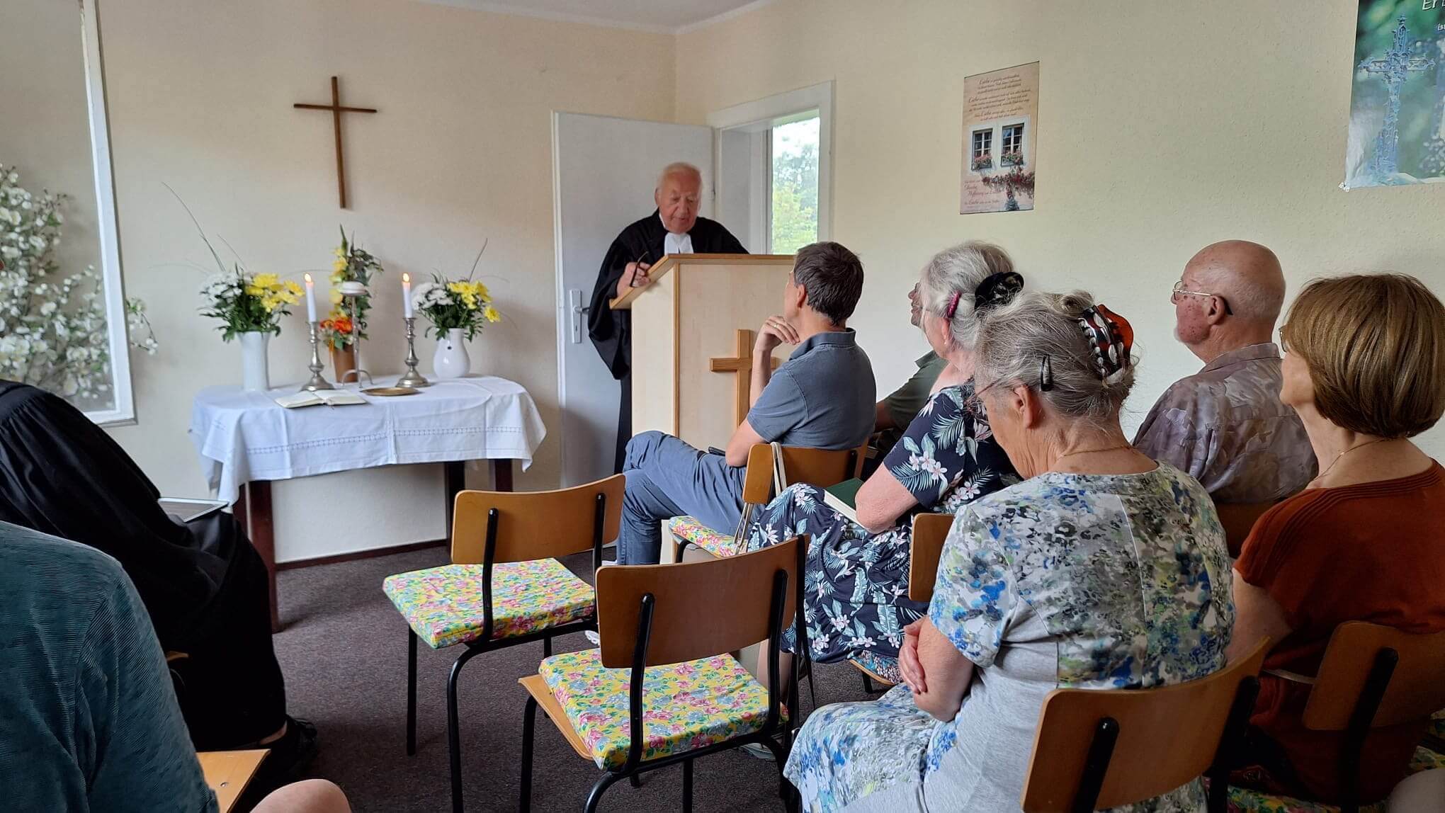 Menschen sitzen in einem kleinen Raum mit Kreuz an der Wand. In der Mitte steht ein Altar mit Kerzen. Ein Pfarrer im Talar steht hinter einem Pult.