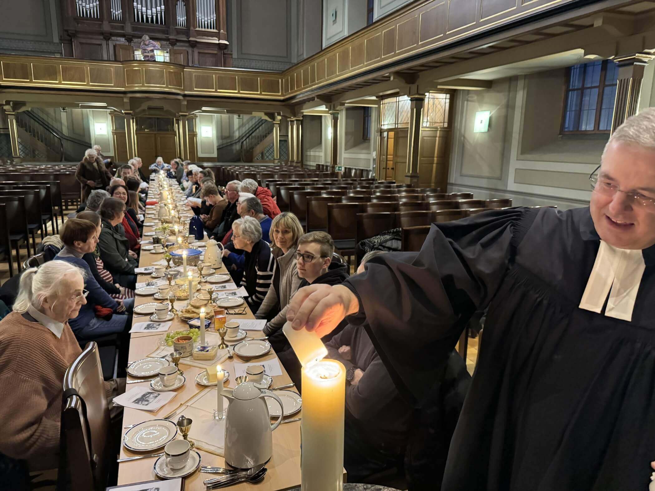 Im Vordergrund zündet ein Pfarrer im Talar eine Kerze an. Im Hintergrund ist eine lange Tafel mit Menschen daran zu sehen. Kerzen leuchten auf der Tafel