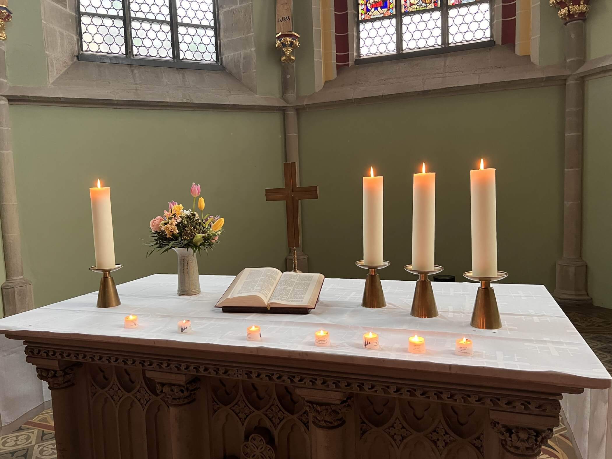 Bild von einem Altar, darauf stehen von links nach rechts: eine Kerze, ein Blumenstrauß, ein Kreuz und drei Kerzen. Davor stehen brennende Teelichter auf dem weißen Tischtuch